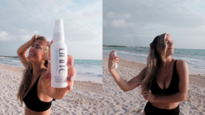 Woman on a sunny beach using and holding up Unite 7 Seconds Detangler spray, smiling with ocean waves in the background.