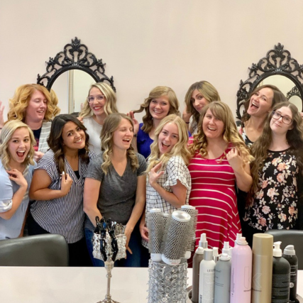 Group of women laughing and smiling together in a salon, gathered around styling stations with hair products displayed in the foreground.