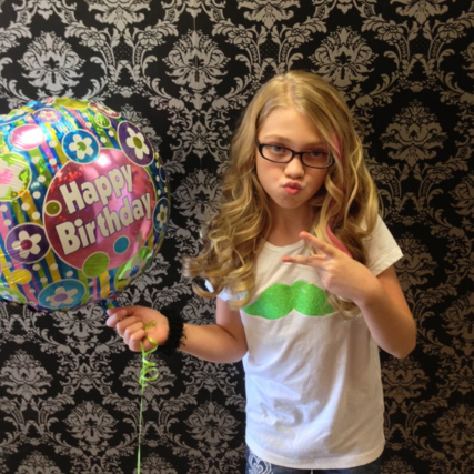 Young girl with long, wavy blonde hair wearing glasses and holding a Happy Birthday balloon inside a salon.
