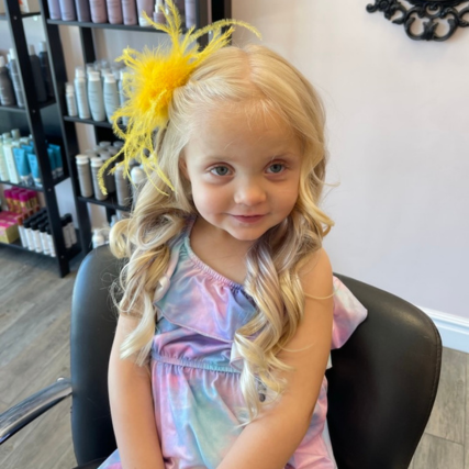 Young girl with long, blonde curled hair and a yellow feather hair accessory, sitting in a salon chair wearing a pastel dress.