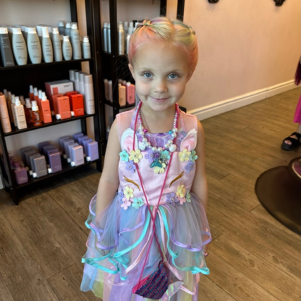 Young girl with colorful braided hair wearing a pastel rainbow dress and flower accessories inside a salon.