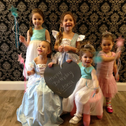 Five young girls celebrating a birthday together in pastel dresses, holding colorful wands and a chalkboard sign that says “Happy Birthday Victoria.”