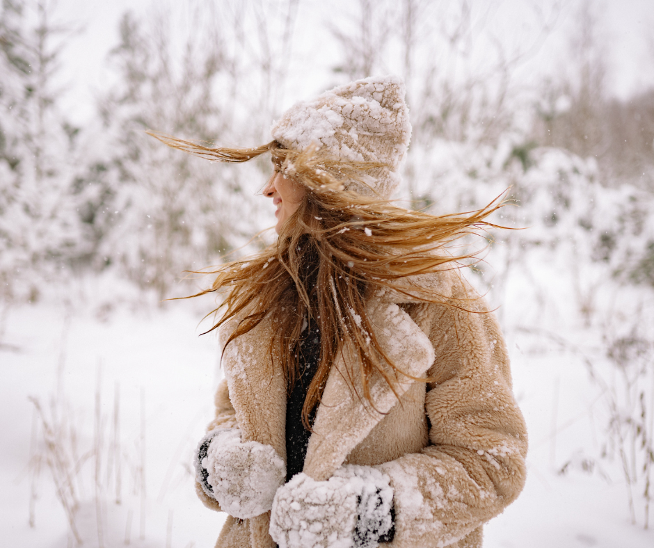 Woman in snowy forest with long hair blowing in the wind, wearing a warm coat and hat