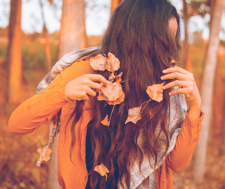 Woman in autumn forest with long hair covered in fall leaves wearing an orange sweater